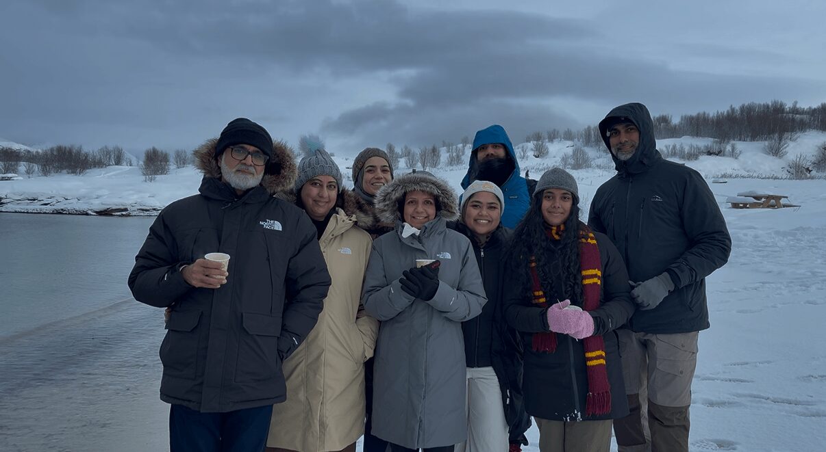 Group of seven people standing outdoors in winter clothing near a snowy landscape and water, cloudy sky.