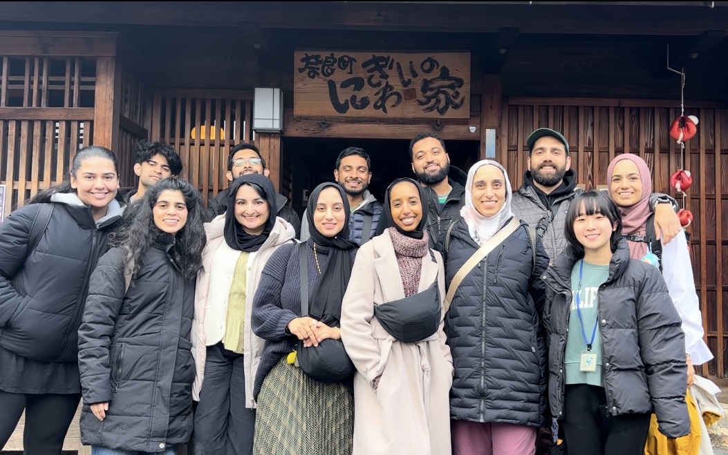 Group of people standing in front of a building with a sign, smiling, wearing winter clothing.