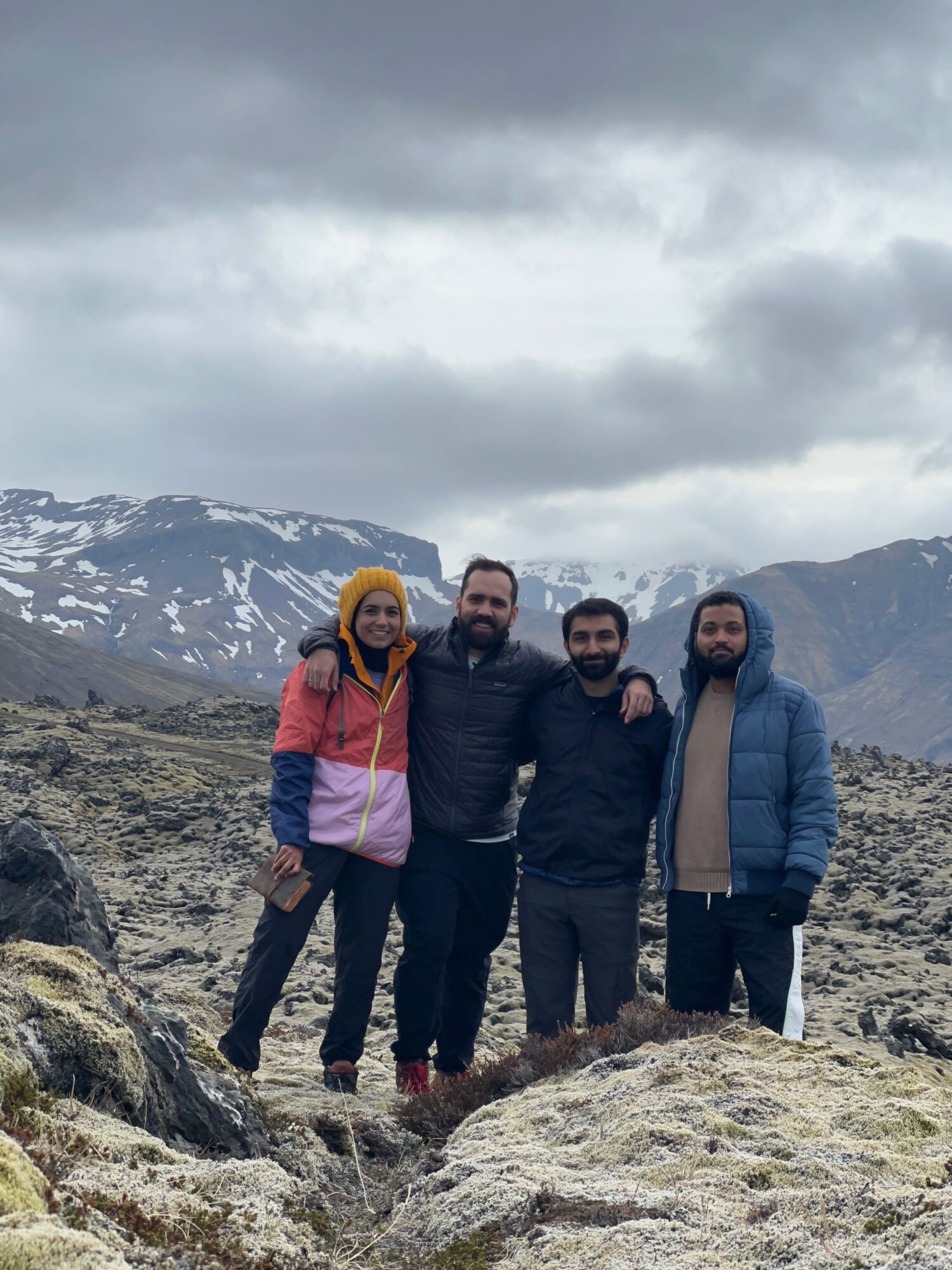 Four people standing outdoors on rocky terrain with mountains and snow in the background, cloudy sky above.