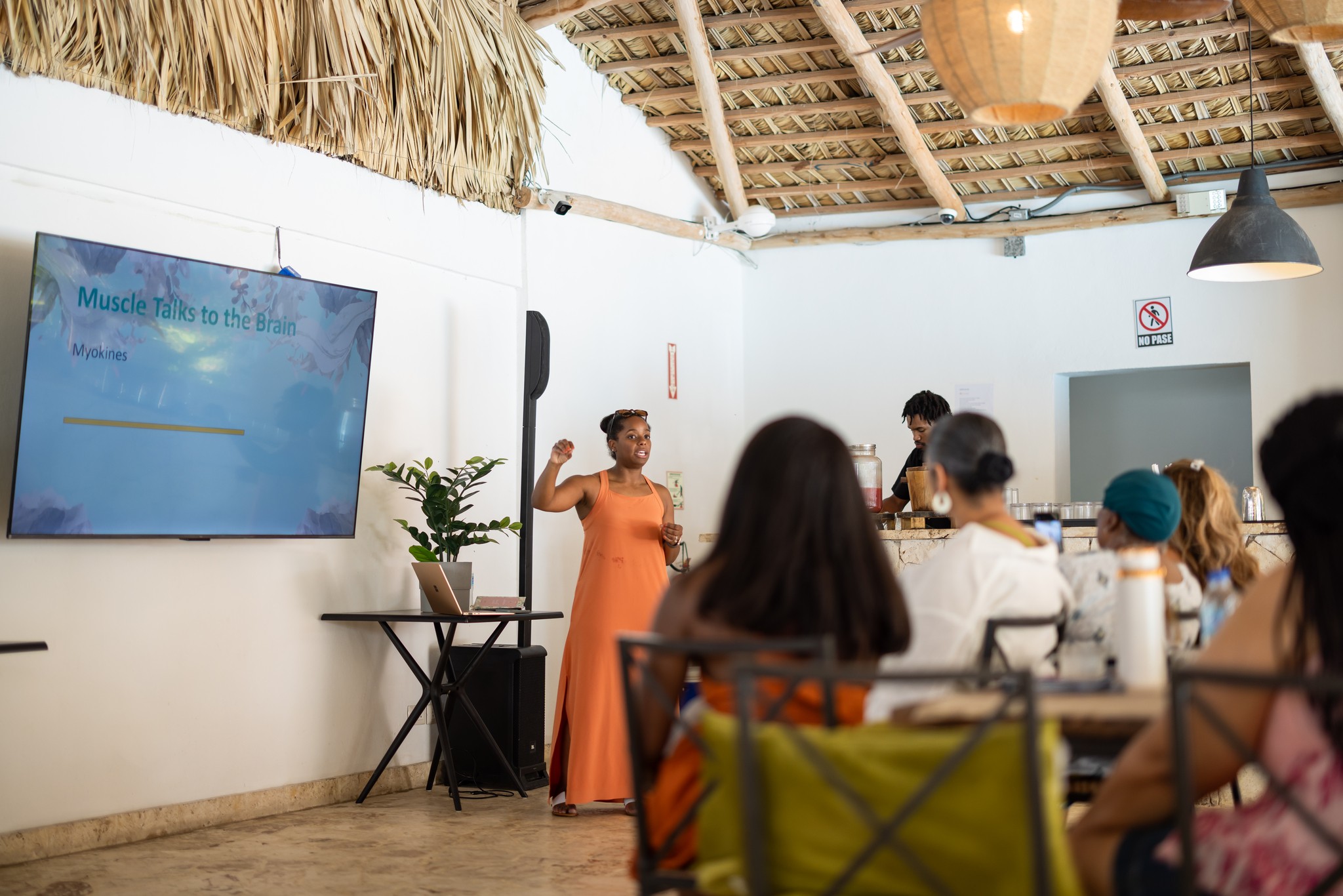 A woman in an orange dress presents to a group in a room with a thatched ceiling and a large screen.