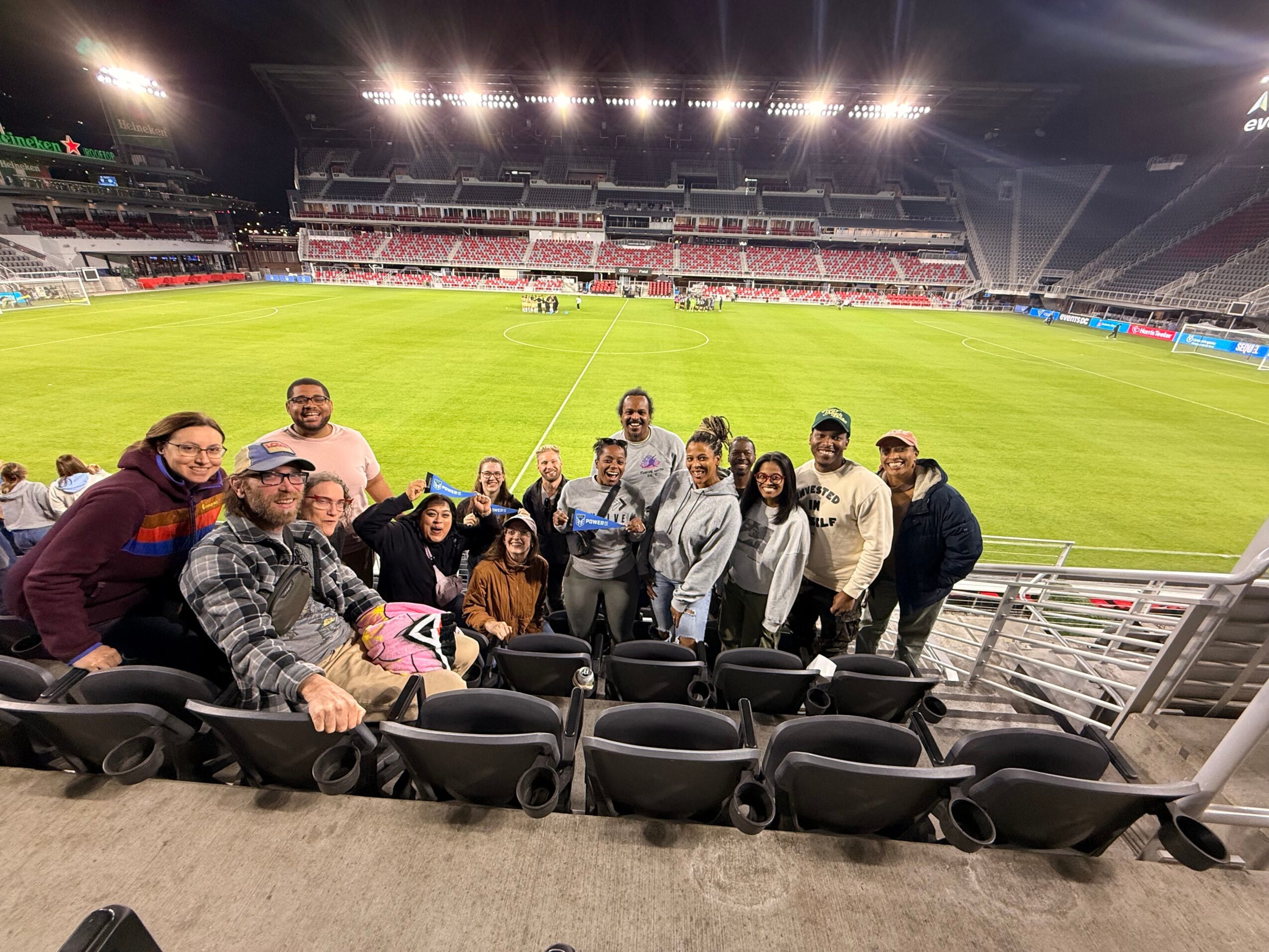 Group of people standing and sitting in stadium seats on a sports field, smiling and posing for a photo.