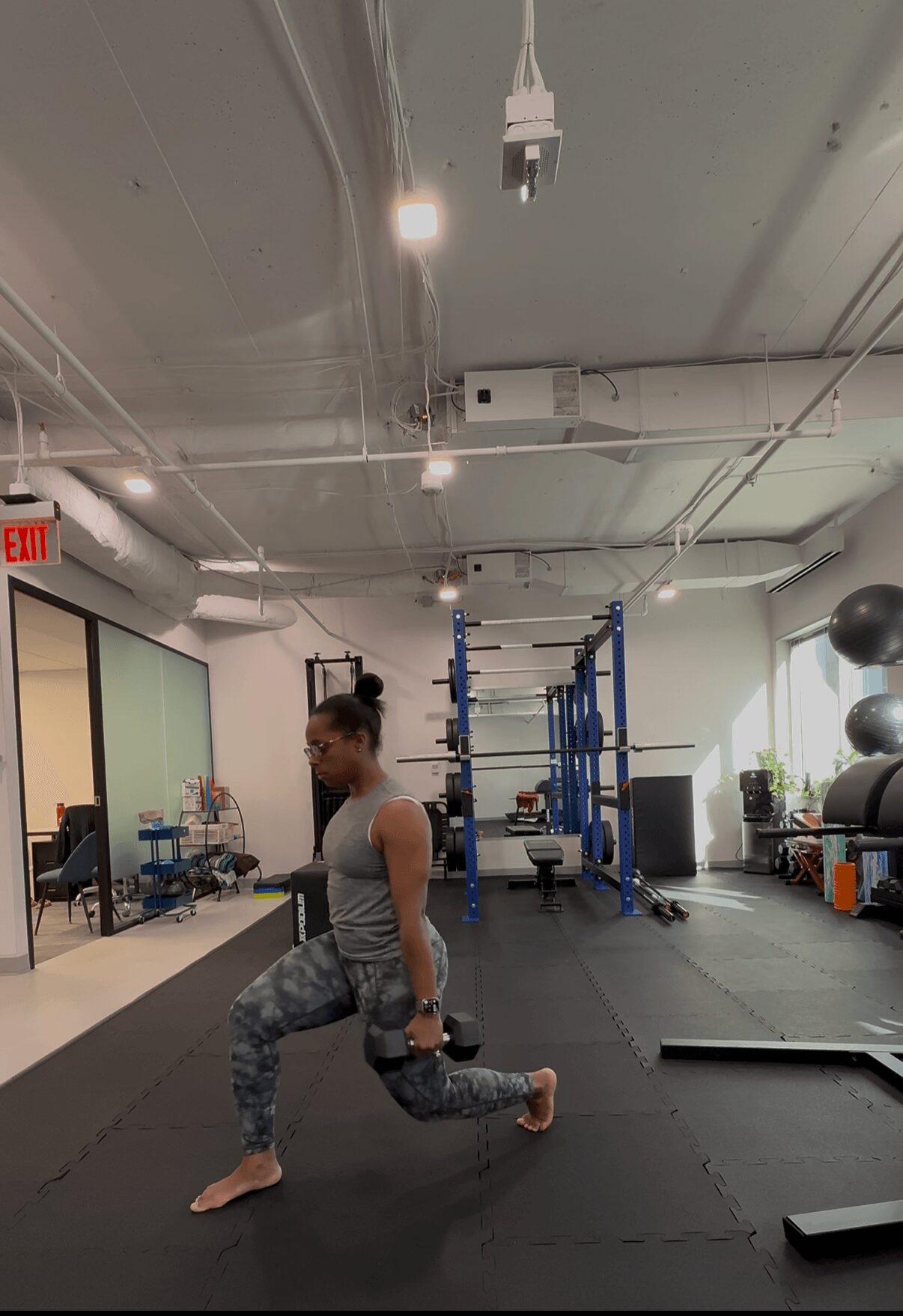 Woman in workout clothes performing a lunge exercise in a gym with equipment and black mats.