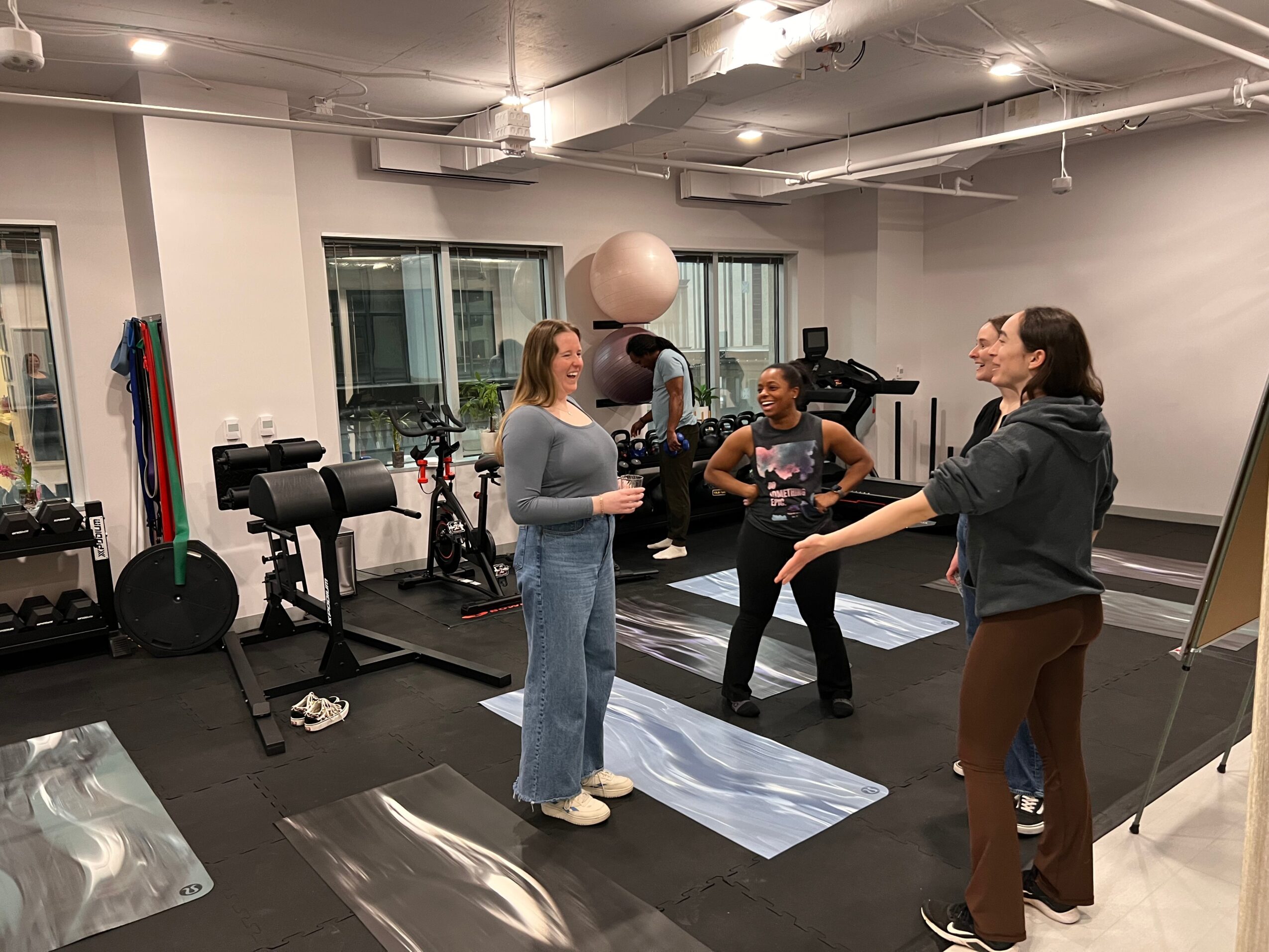 Four women in a gym, one woman speaking, others listening, exercise mats on the floor, gym equipment in background.