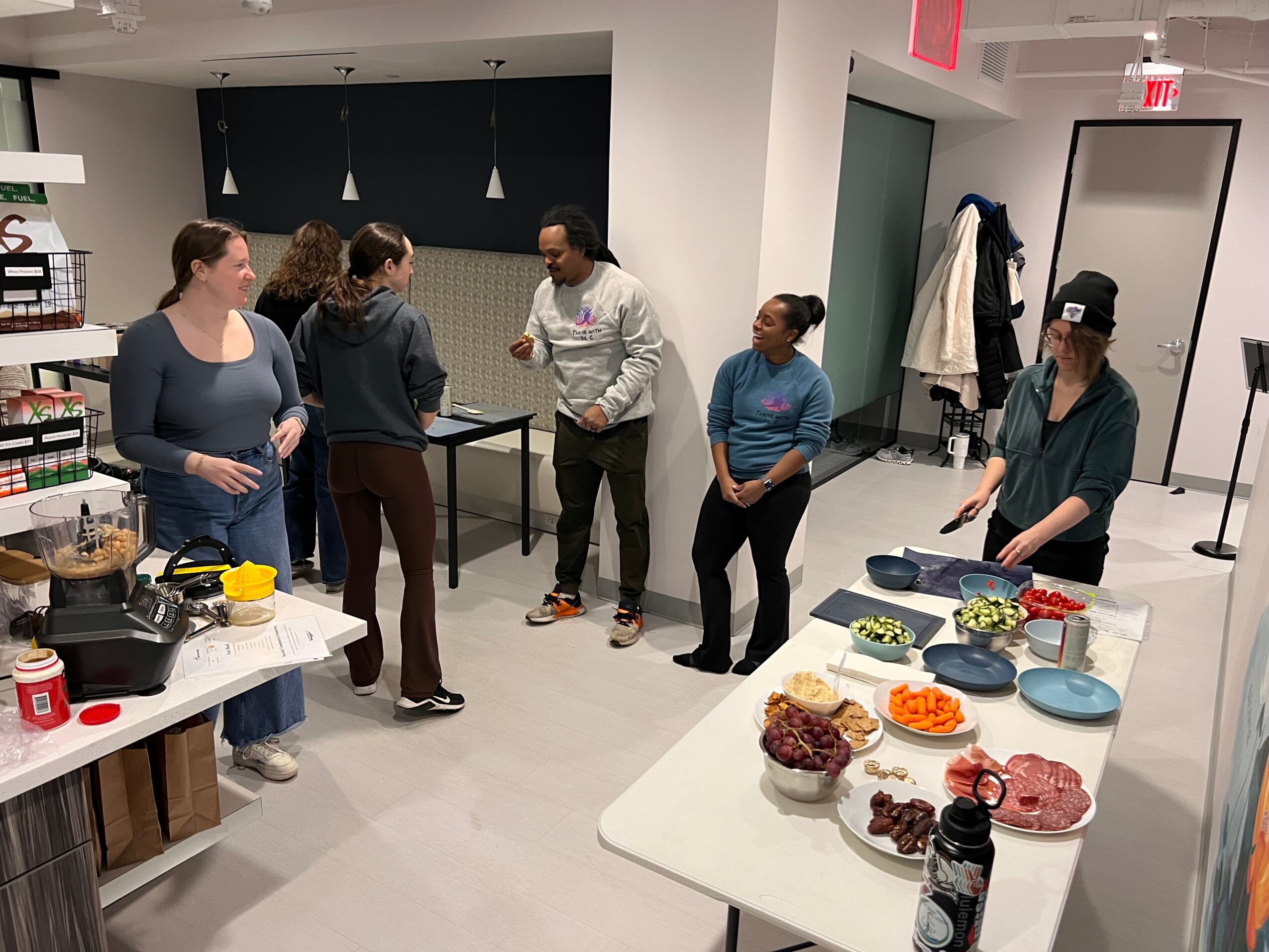 Group of six people standing and talking in a room with a table of food and drinks, and a table with supplies.