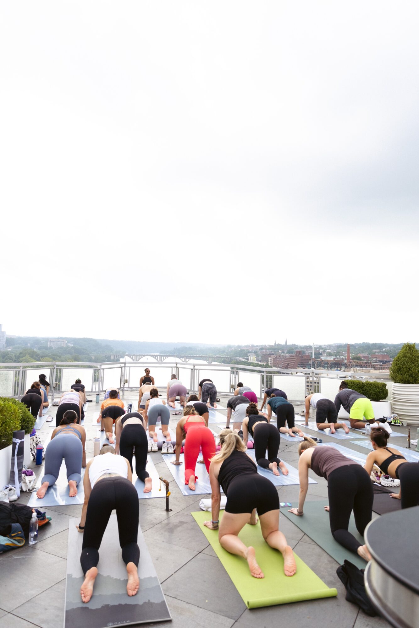 People practicing yoga outdoors on a rooftop with a city view, some in downward dog pose, overcast sky.