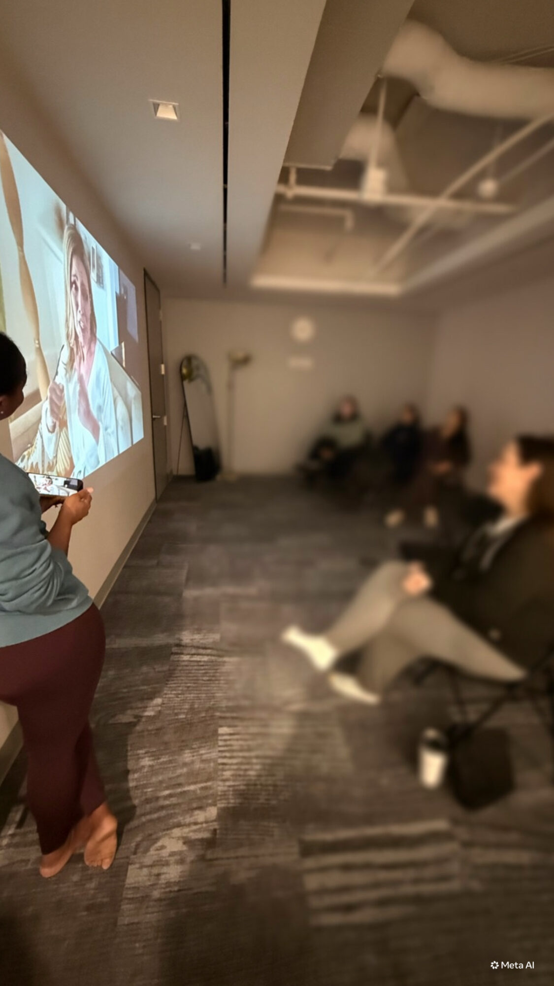 Person standing and speaking to seated group in a room with a large screen displaying a video call.