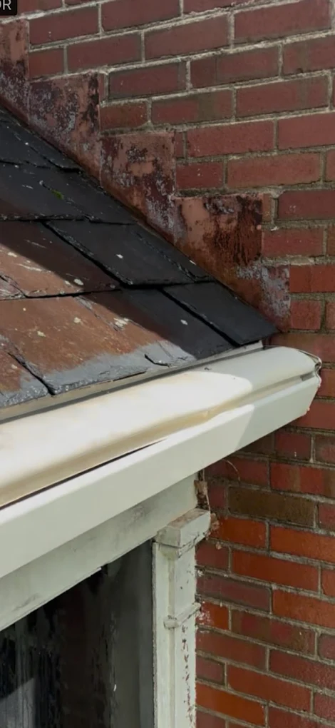 Close-up of a brick wall and roof edge with black shingles and white gutter, showing weathered bricks and roof details.