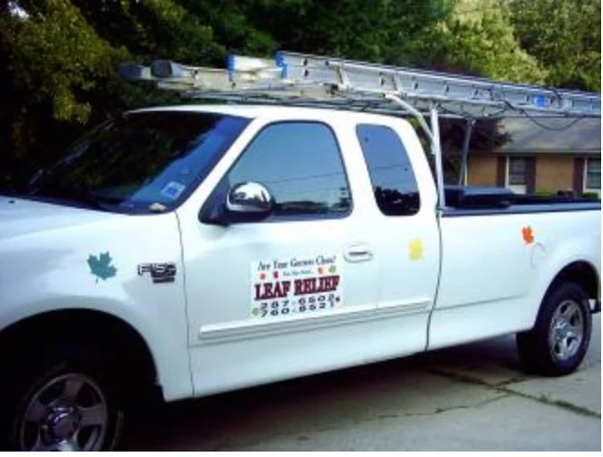 White utility truck with ladder on top, parked outdoors with trees and a building in the background, leaf decals on the side.