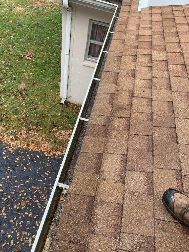 View from roof showing shingles, a person's shoe, and a house with a window, with grass and driveway below.