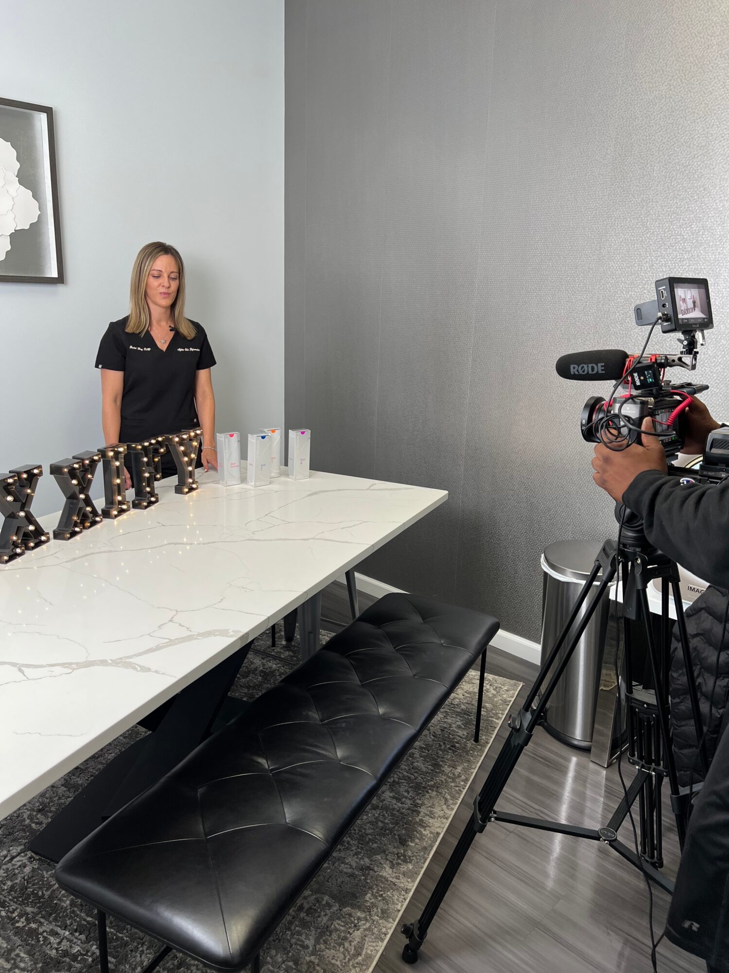 Woman standing near a white table with glasses, camera operator filming her in a room with gray walls.