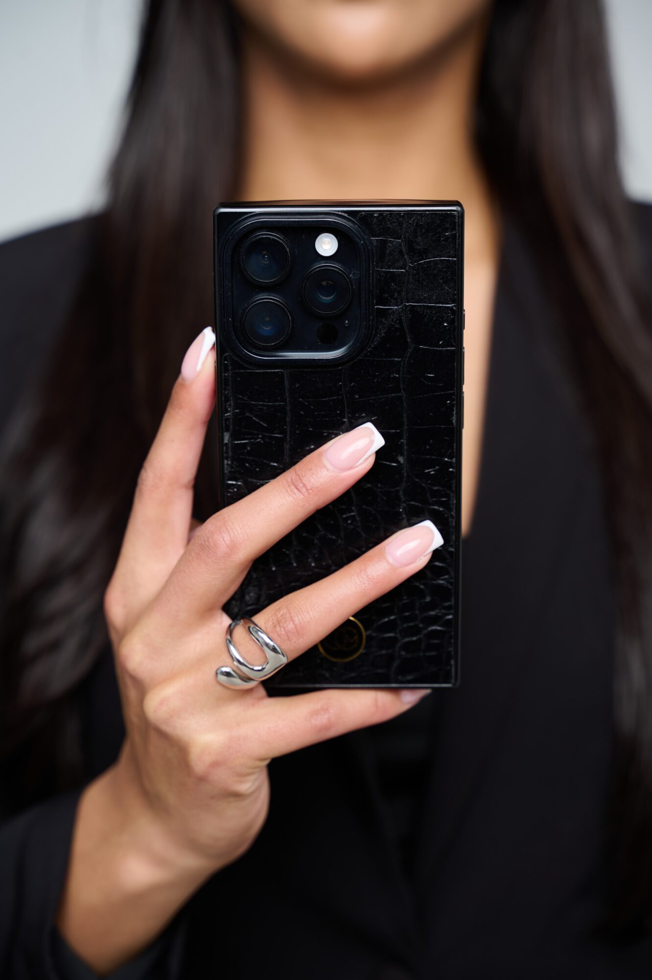 Person holding a black smartphone with a textured back, fingers visible, face partially shown, dark hair, black clothing.