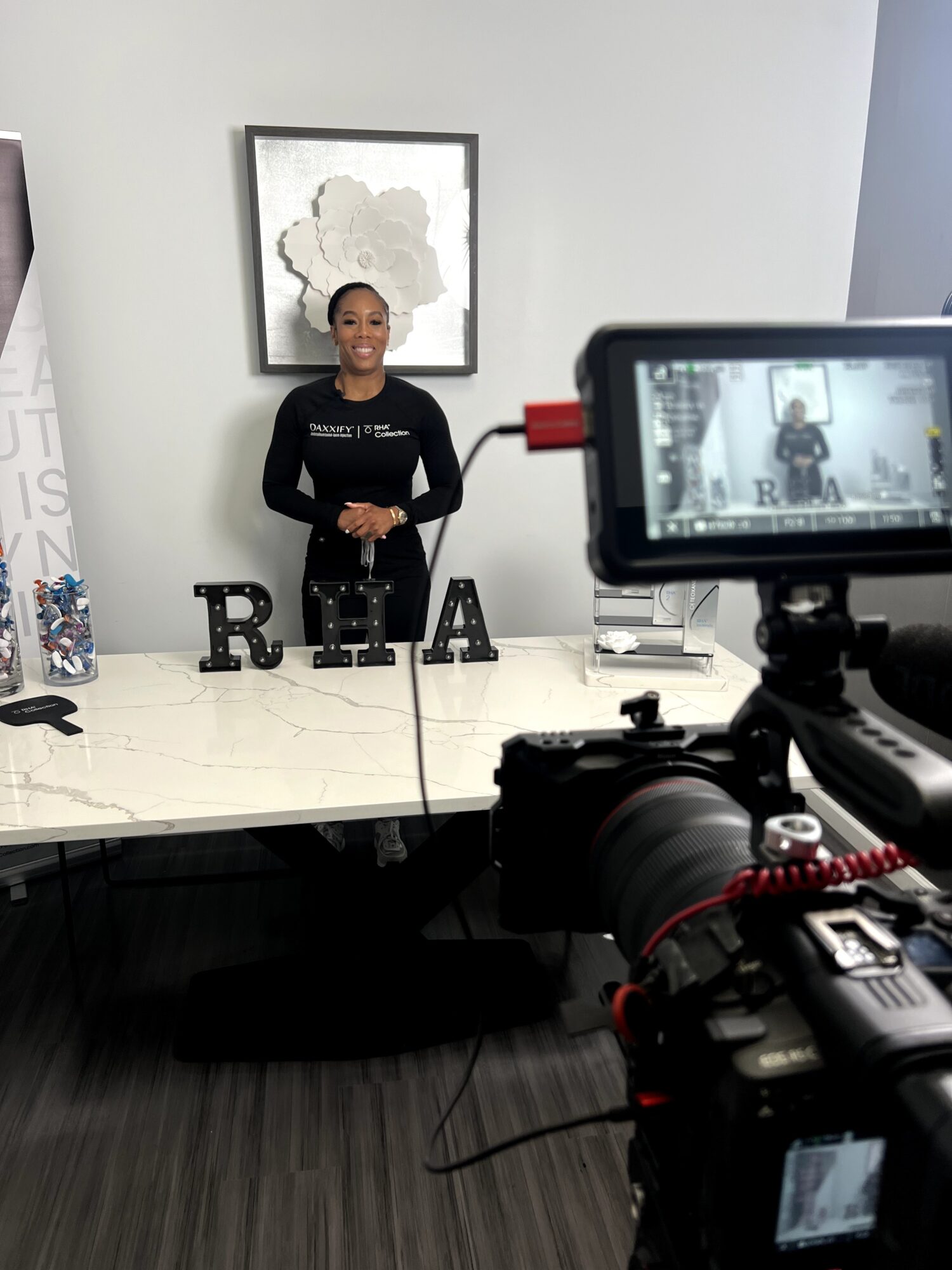 Woman standing behind a table with RHA letters, camera recording her, abstract art on wall, neutral background.