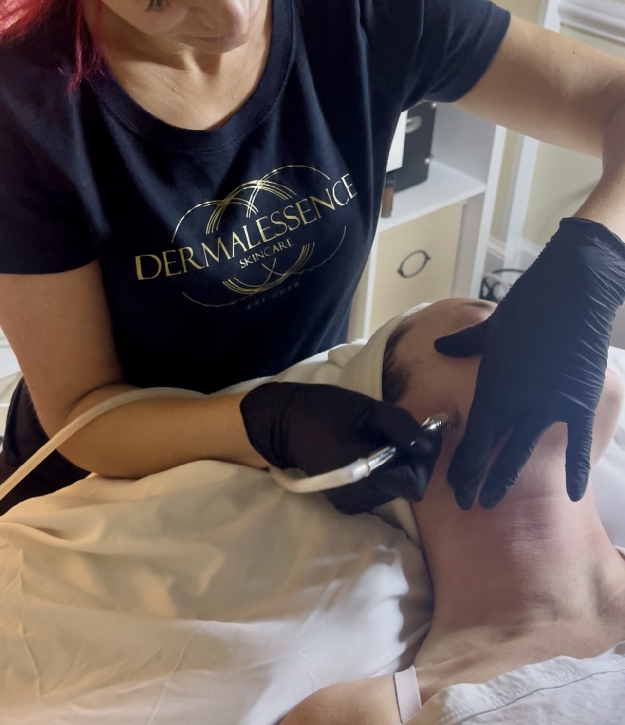 Person receiving a facial treatment, lying on a bed, with a practitioner wearing black gloves applying a device to the face.