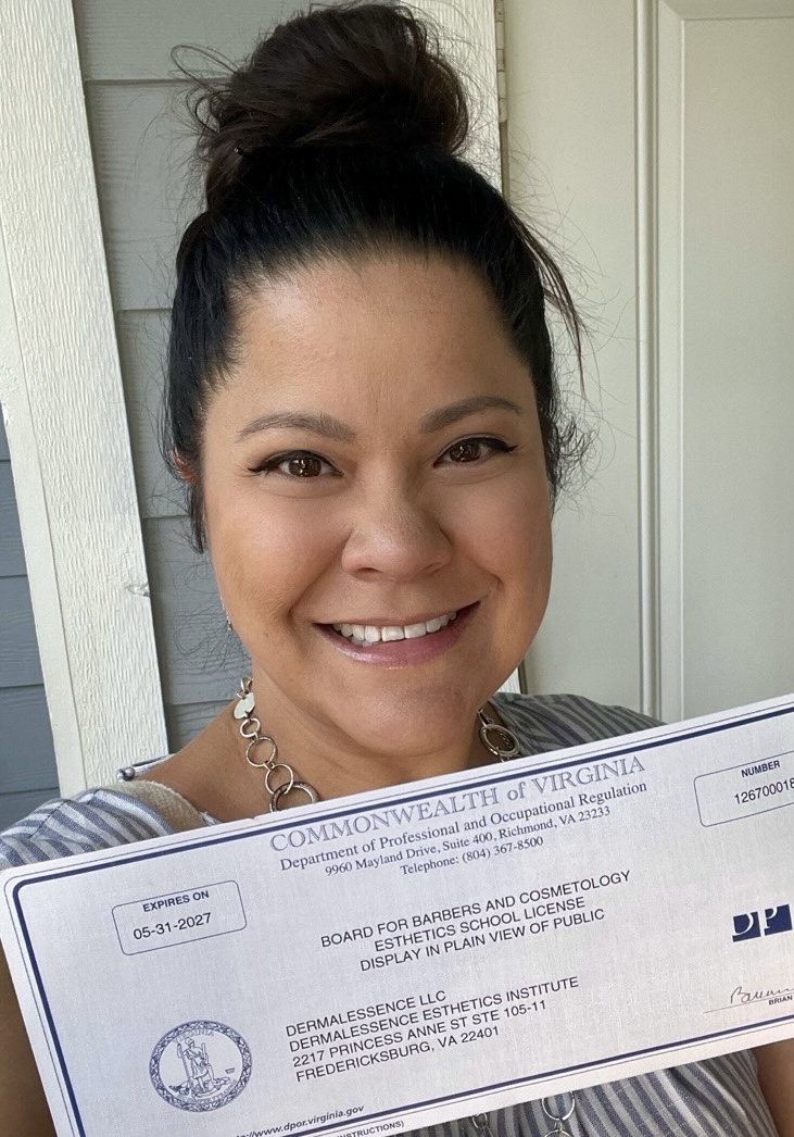 Smiling woman with dark hair in a bun holding a certificate, standing outside near a door.