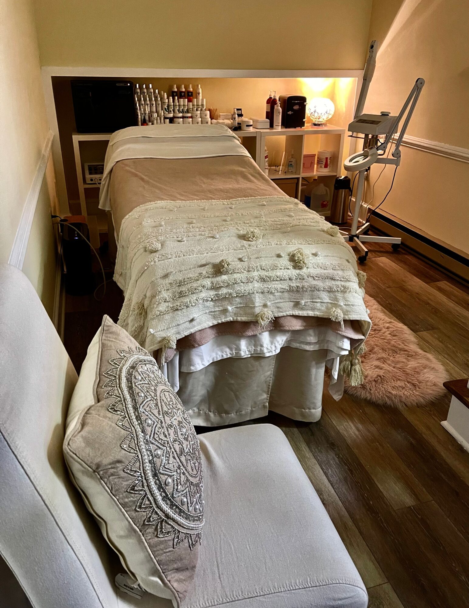 A cozy room with a bed covered in a beige blanket, a pillow with intricate embroidery, and a wooden floor.