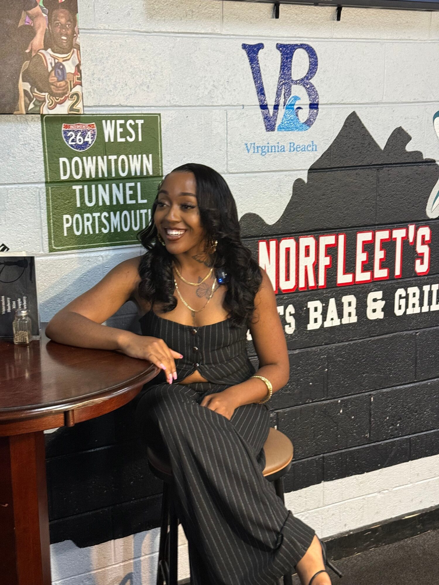 Woman with curly hair playing guitar, sitting at a table, smiling, in front of a wall with signs and posters.