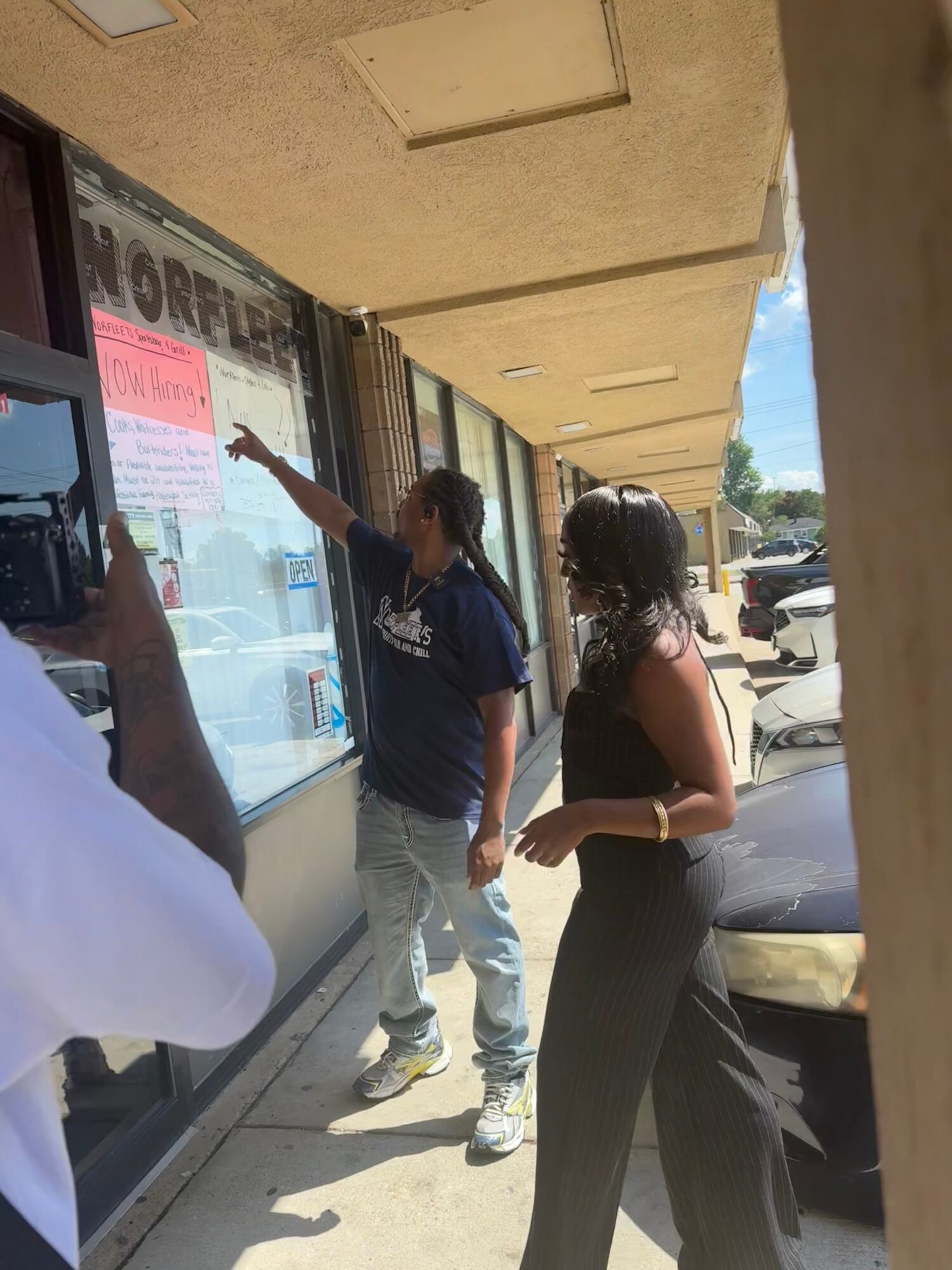 Two women stand outside a storefront window, one pointing at a sign, with a person taking a photo nearby.