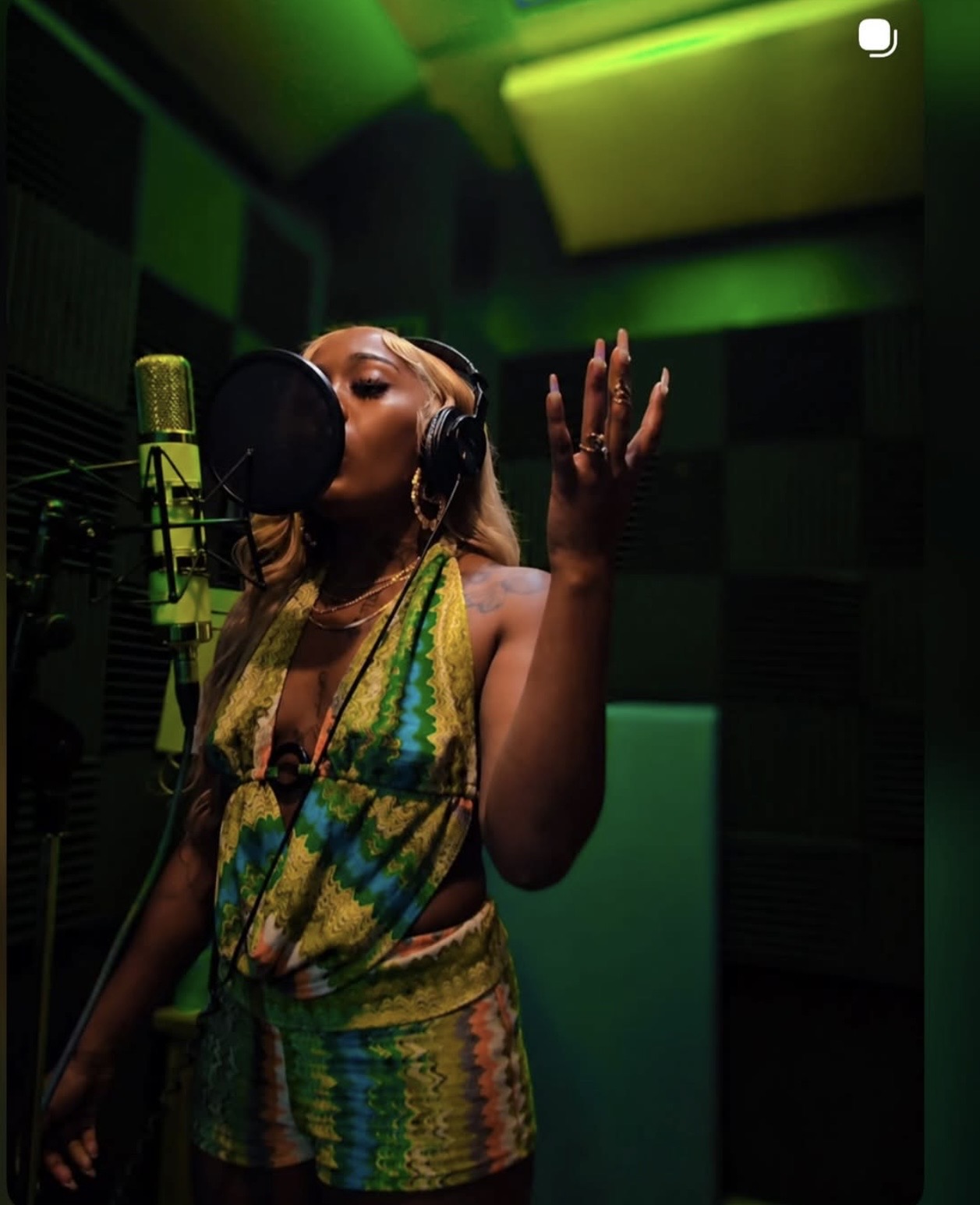 Woman singing into a microphone in a recording studio with green and yellow lighting, wearing a colorful striped top and headphones.