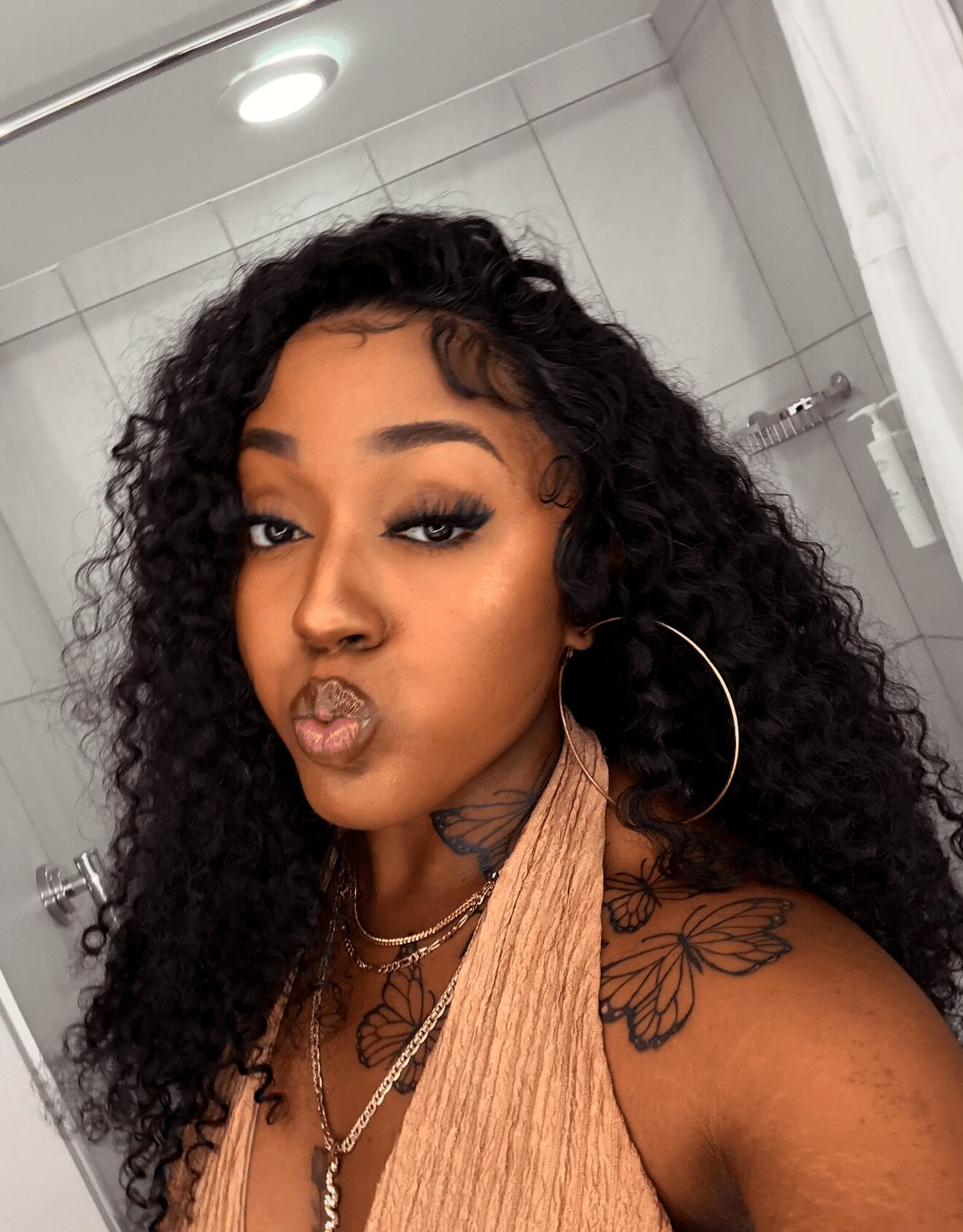 Woman with curly hair making a kiss face in a bathroom, wearing hoop earrings and a beige top, with tattoos visible on shoulder.