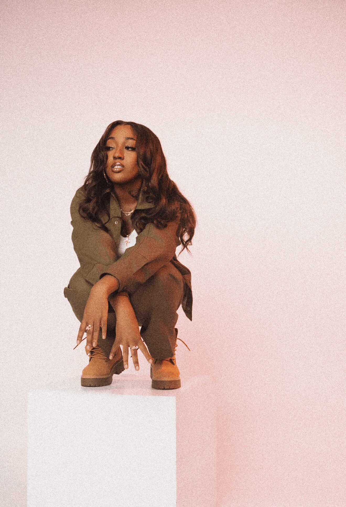 Young woman crouching on a white block against a pink background, wearing a brown outfit and boots.