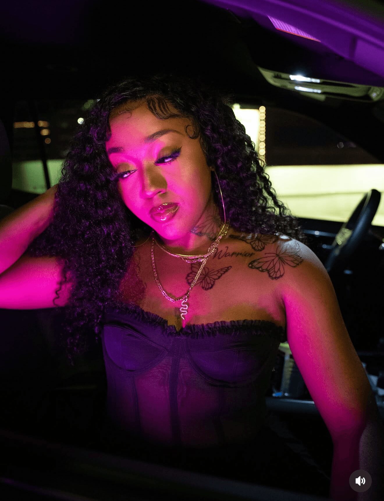 Young woman with curly hair in a car, illuminated by pink and purple lights, wearing a strapless top and jewelry.