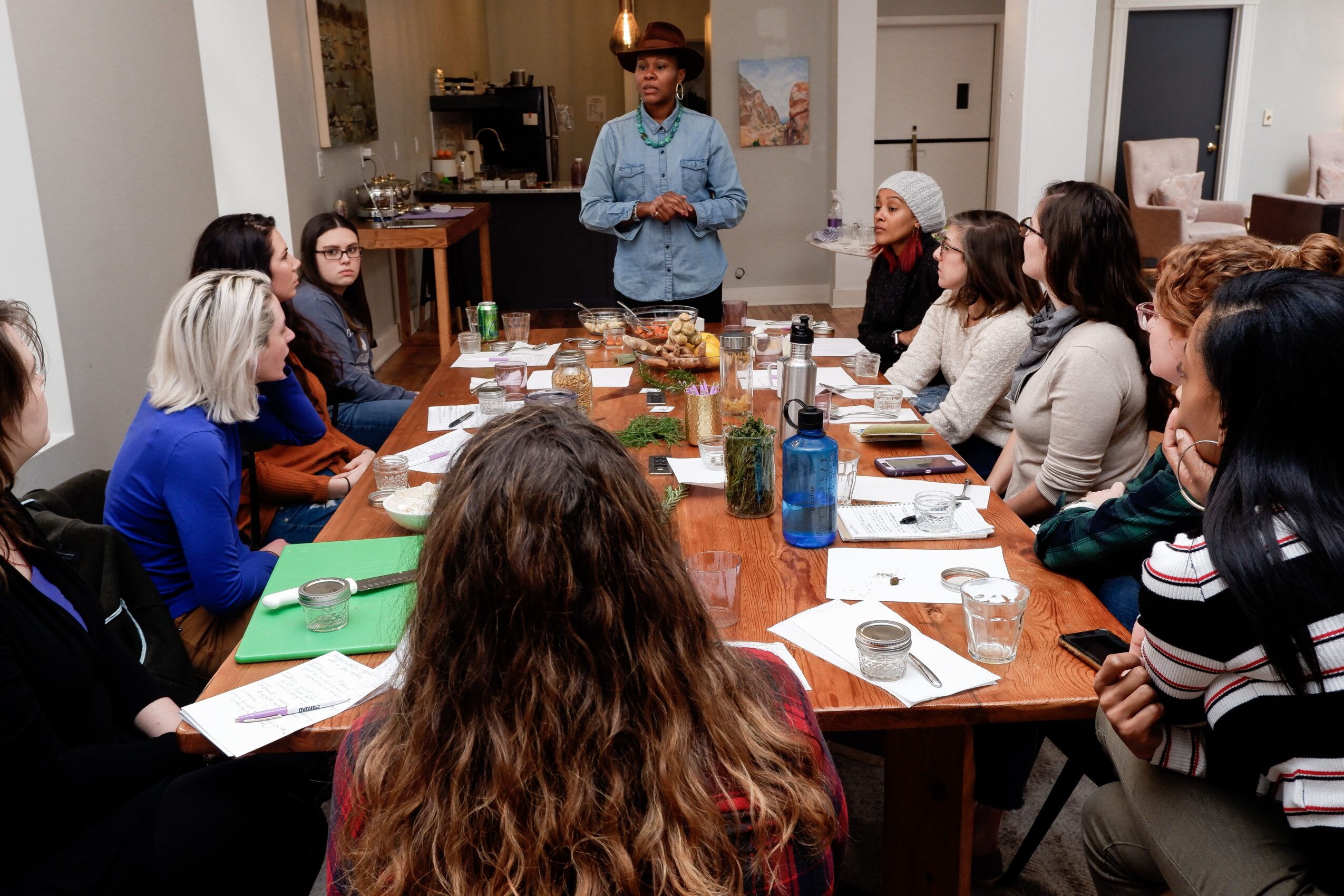 Group of people seated around a table listening to a person standing and speaking in a room, with notebooks and drinks on the table.