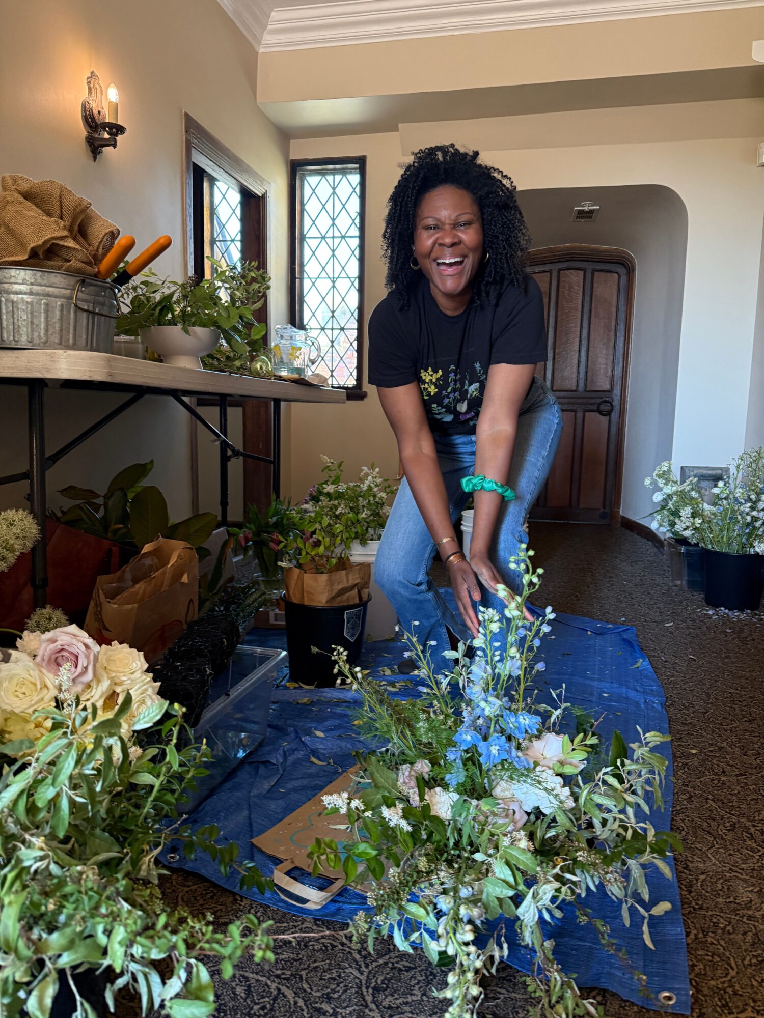 Woman with curly hair in black top and blue jeans, smiling, standing beside a floral arrangement on a table covered with a blue cloth, in a room with a window and wooden furniture.