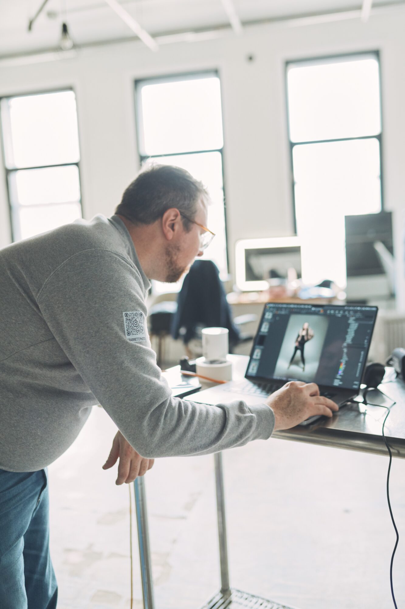 Man working on a computer in a bright, modern office with large windows, using photo editing software.