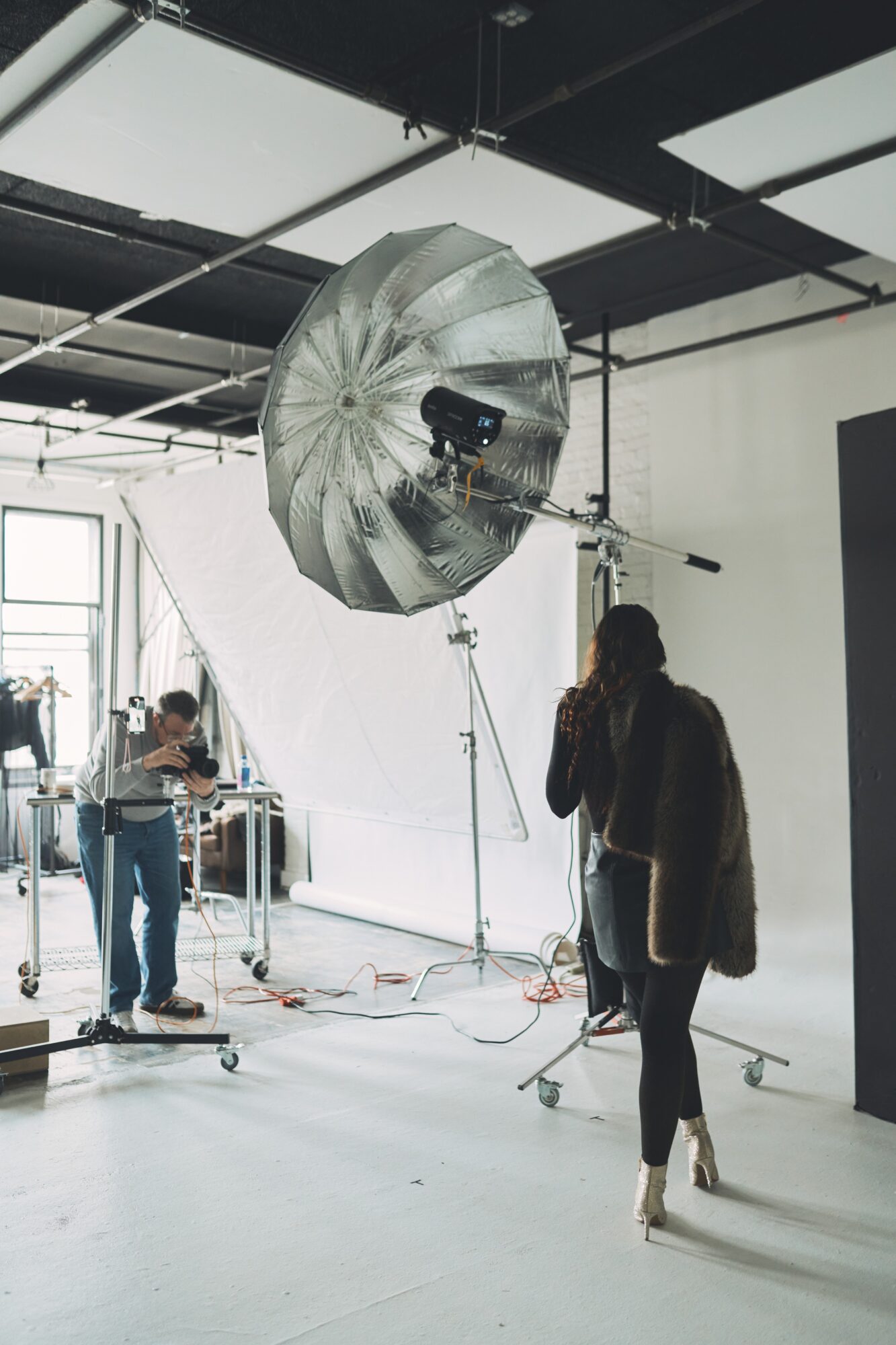 Person standing in a photography studio with large reflective umbrella and equipment, bright window in background.