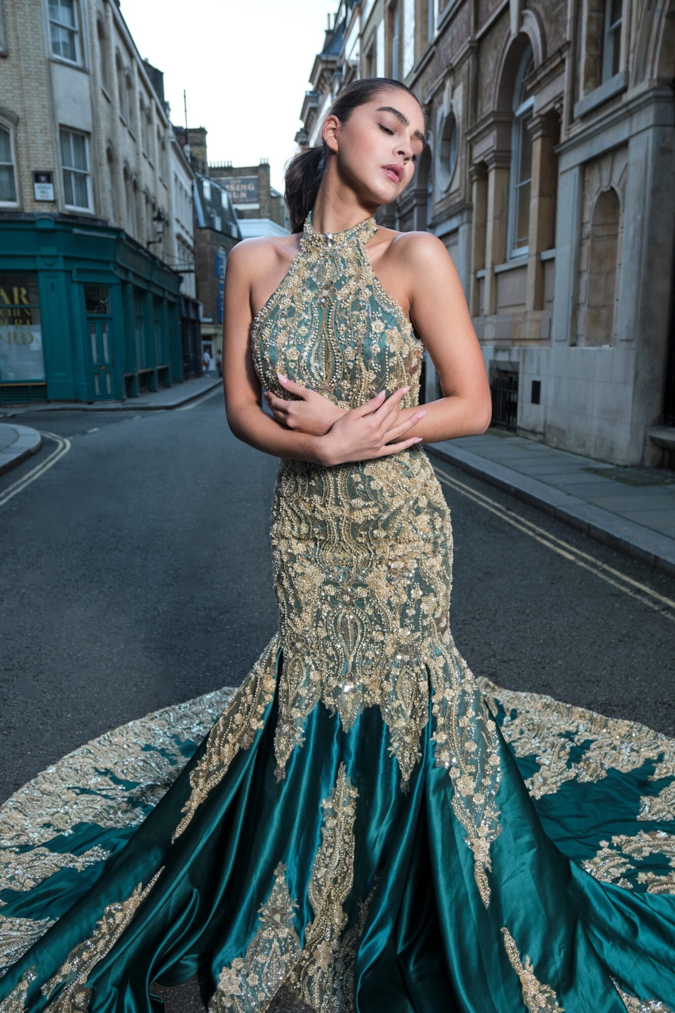 Woman in ornate gown standing on city street with buildings in background, arms crossed, eyes closed, elegant pose.
