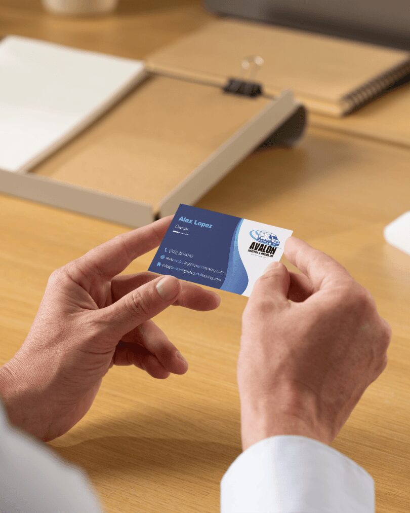 Person holding a business card with both hands over a wooden desk, with notebooks and papers in the background.