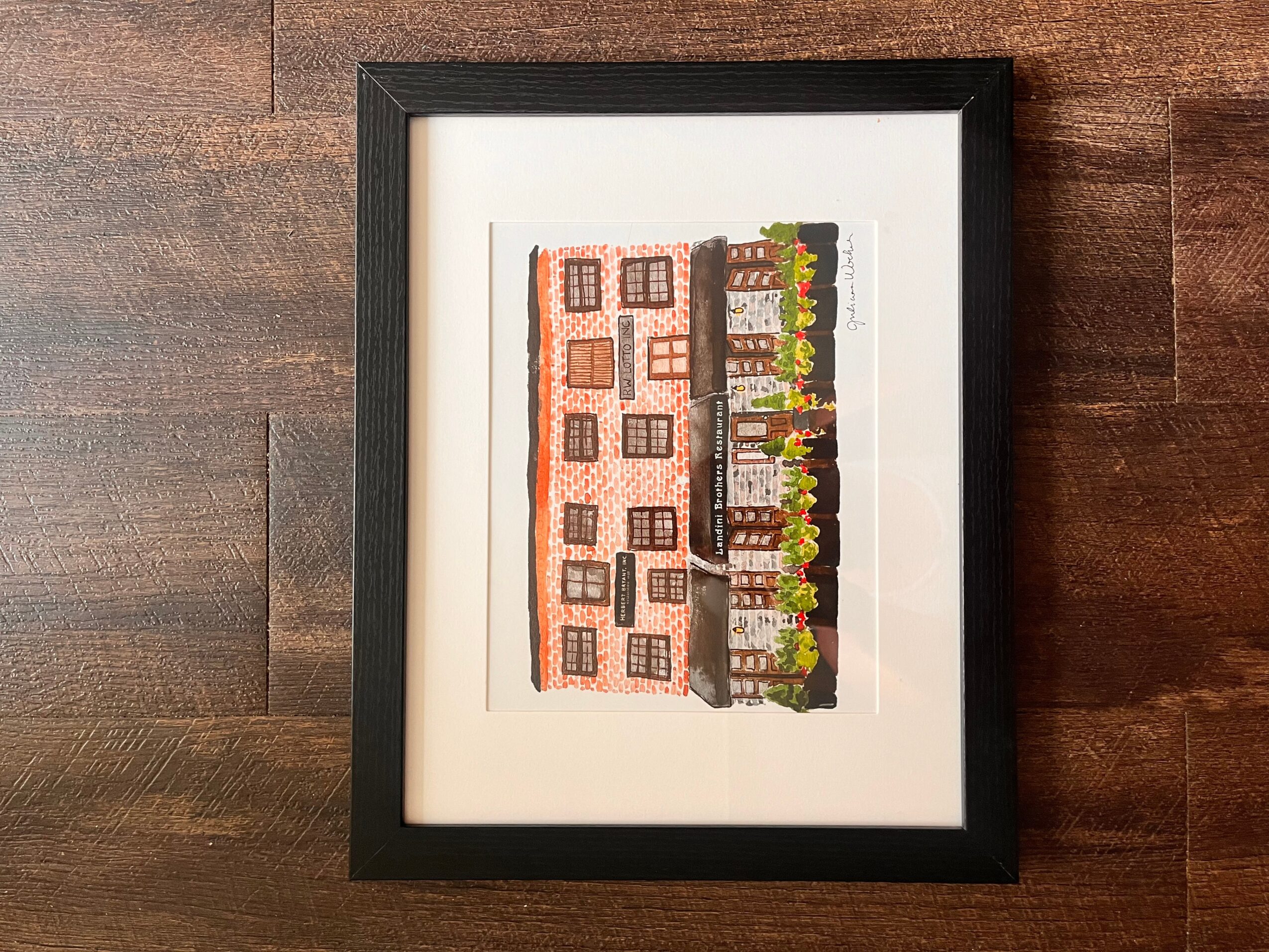 Framed colorful illustration of a building with windows and a garden, on a wooden surface.