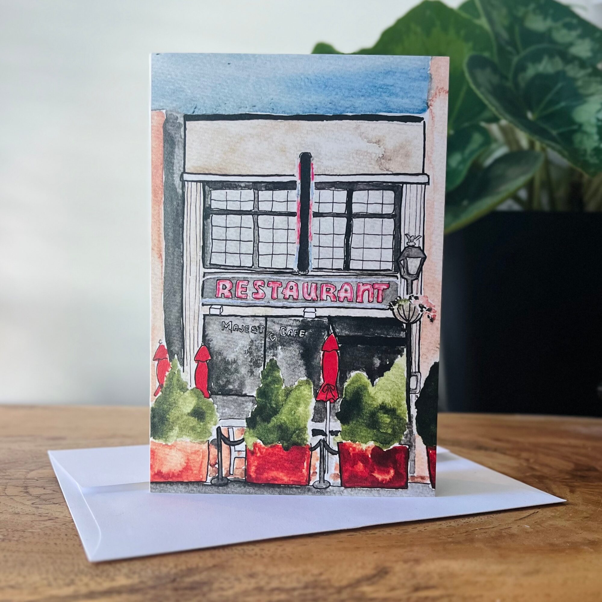 Watercolor painting of a restaurant facade with a sign, windows, and greenery in front, on a wooden surface with a plant in background.