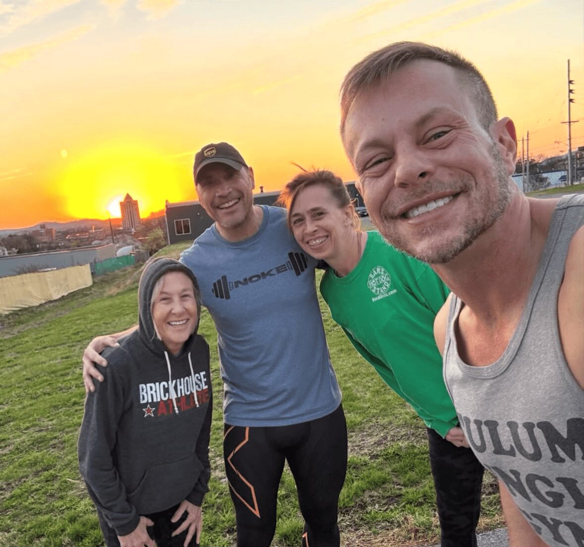 Four people smiling outdoors at sunset, standing on grass with buildings and power lines in background.