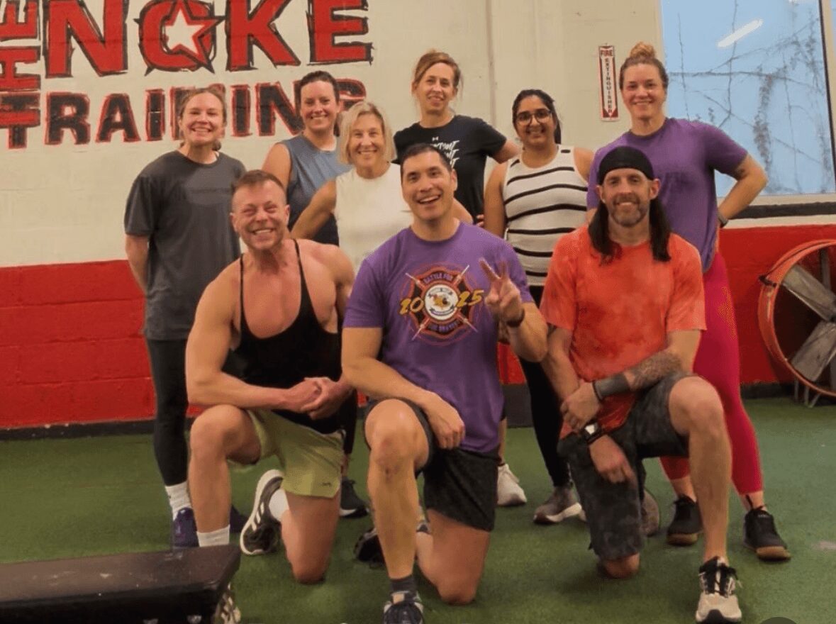 Group of ten people posing inside a gym with a red and white wall and a large wheel in the background.