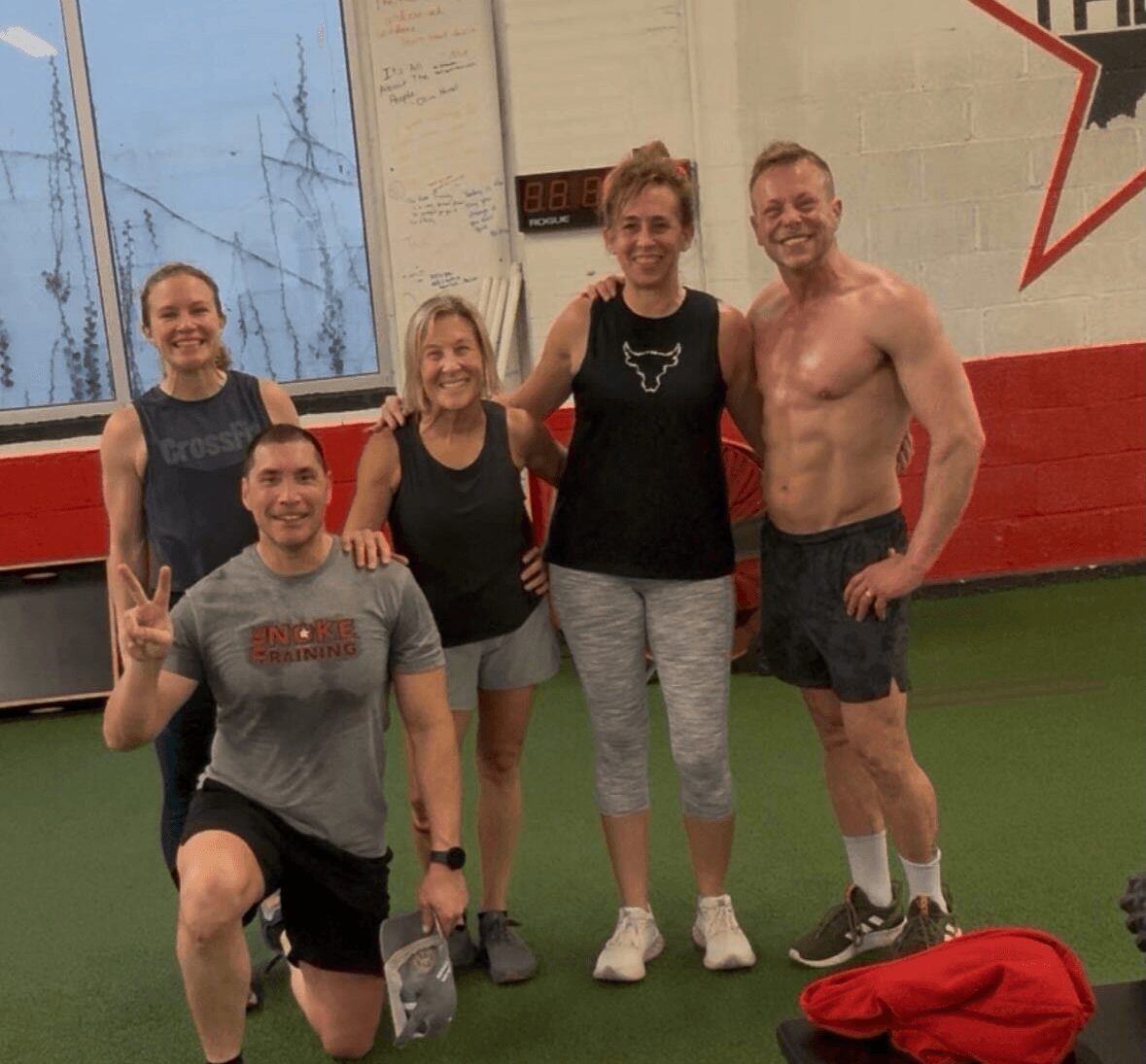 Group of five people smiling indoors, one person making a peace sign, in a gym setting with a red and white wall.