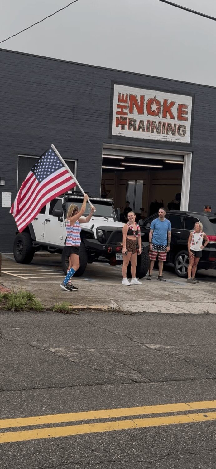 Group of people standing outside a building with a sign that reads 'The Noke Training,' one person holding an American flag.