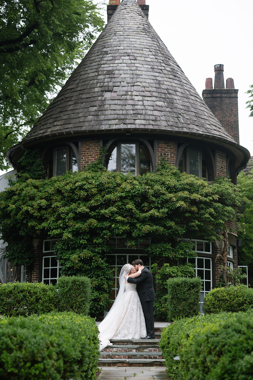 A bride and groom embrace in front of a round, turreted brick house with a conical roof, surrounded by greenery.
