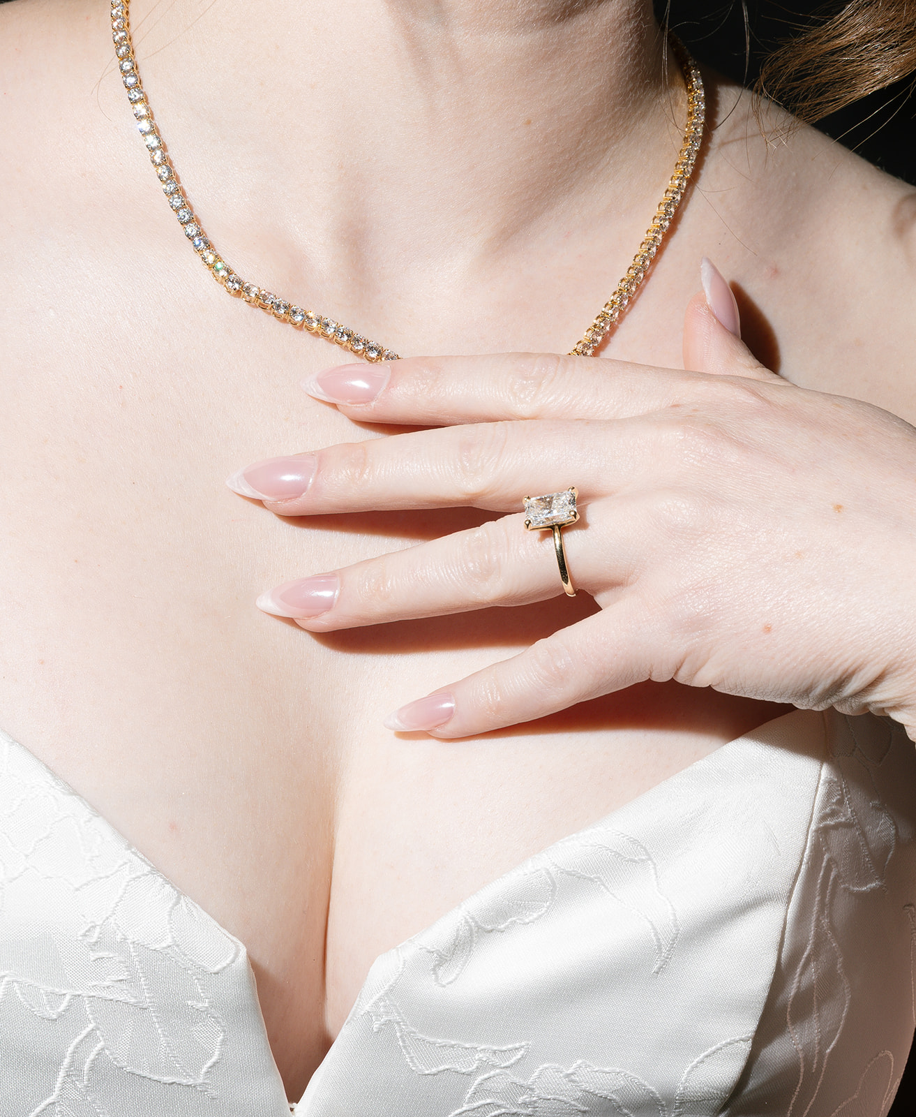 Close-up of a woman wearing a gold necklace and a ring, with her hand resting on her chest, in a white top.