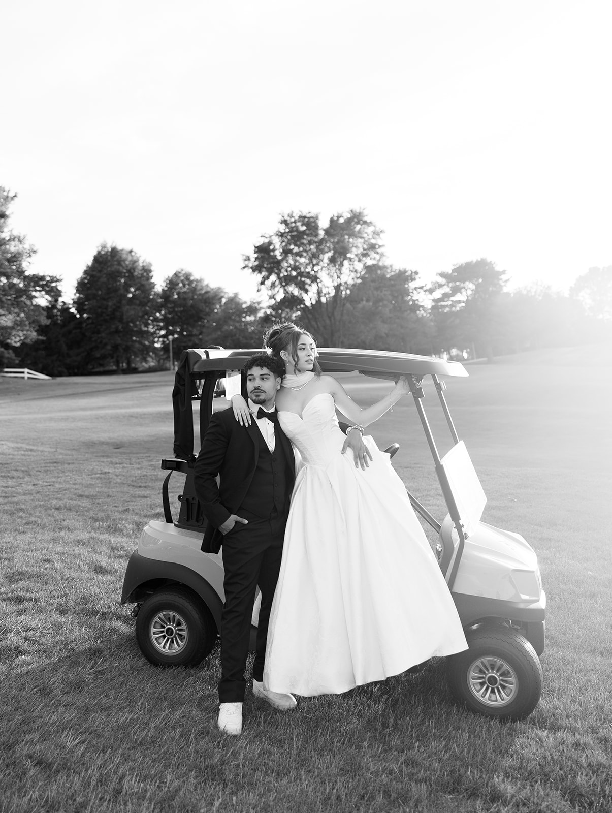 A couple in formal attire standing next to a golf cart on a grassy field with trees in the background.