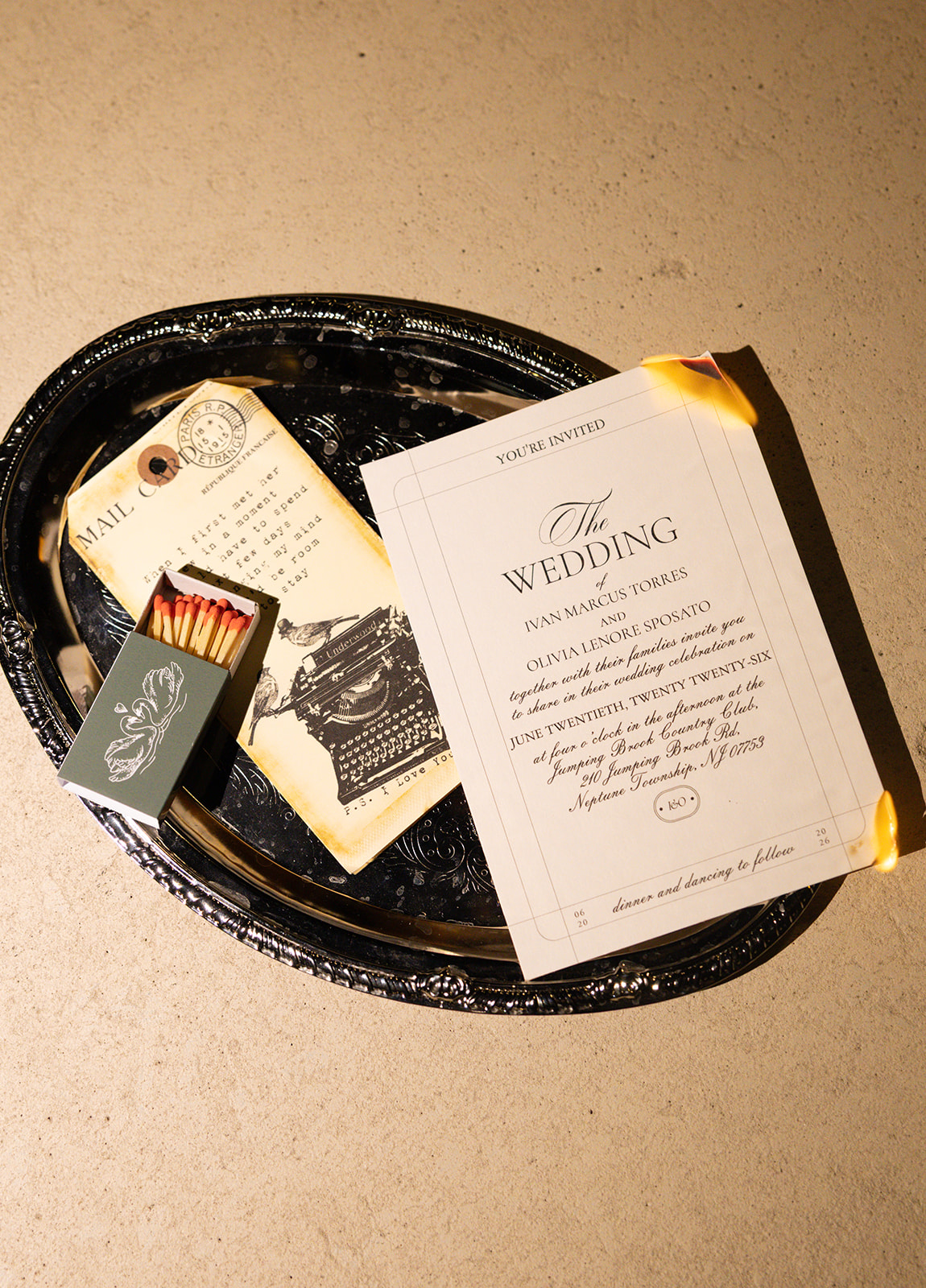 A black tray holds wedding invitations, a matchbox, and a small card on a beige surface.