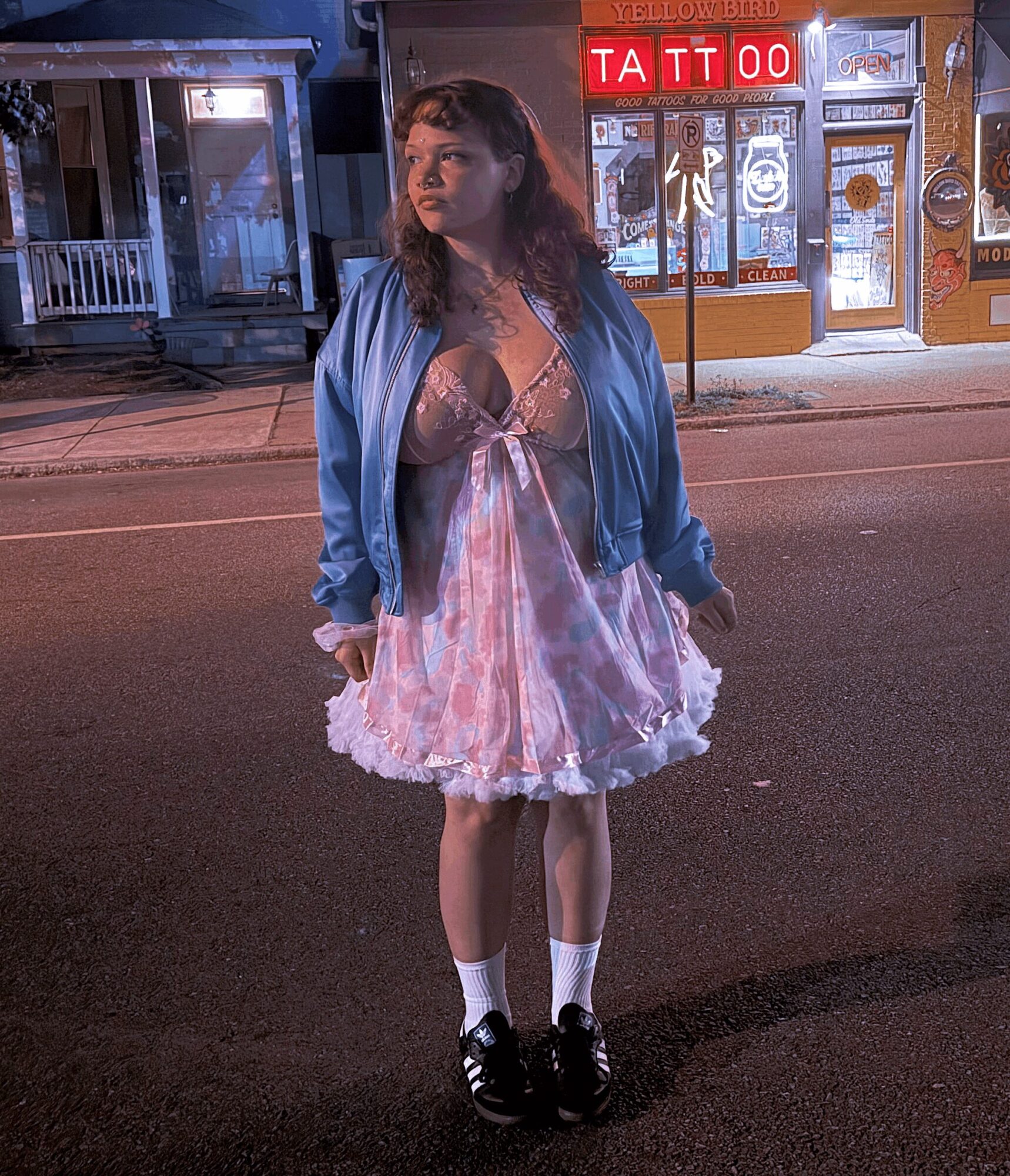 Young woman standing on street at night, wearing a pink dress, blue jacket, and black shoes, with storefronts in background.
