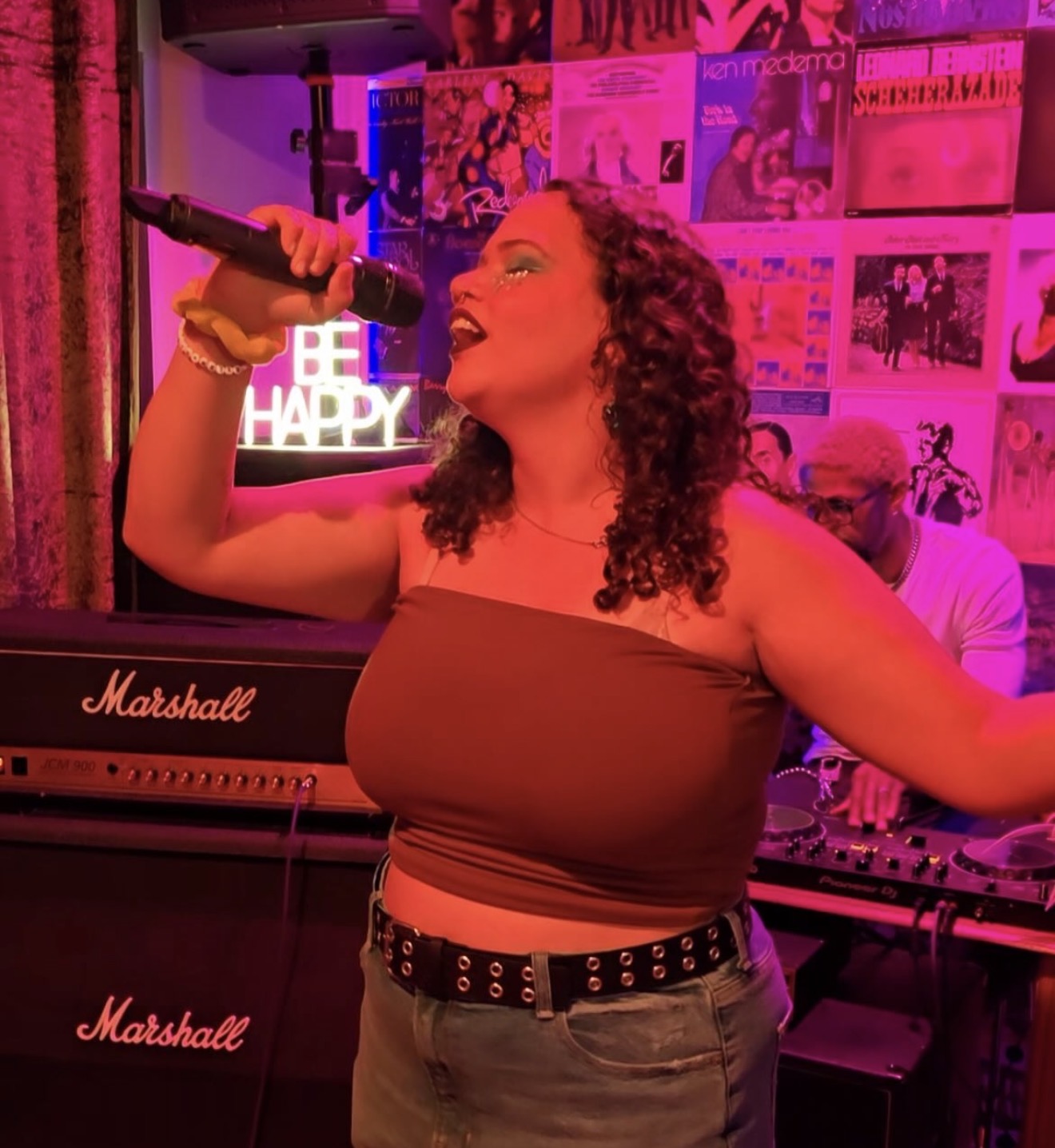 Woman singing into microphone in a music venue with pink lighting and posters on the wall.