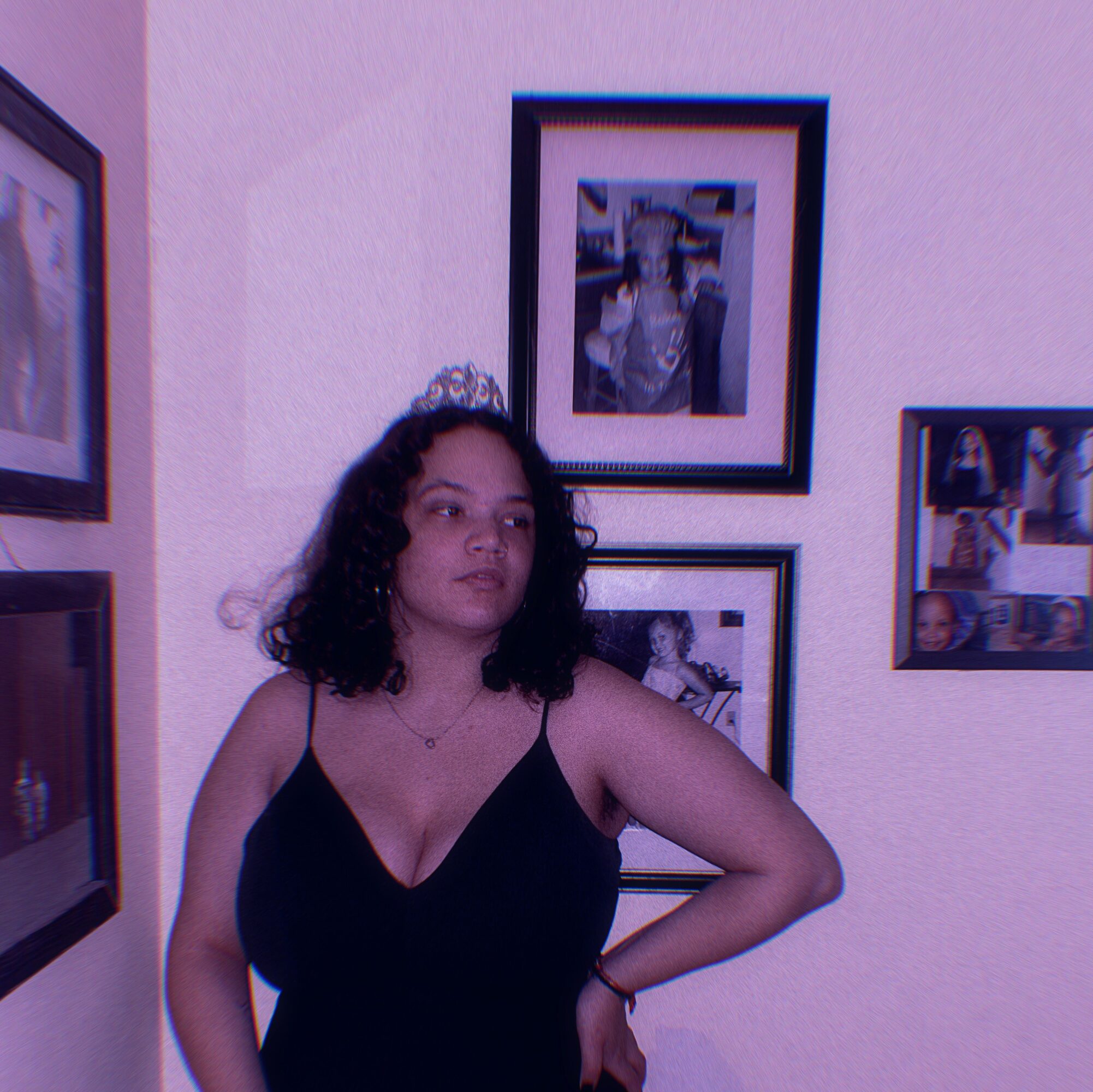 Young woman with curly hair in a black dress standing in front of framed photos on a wall.