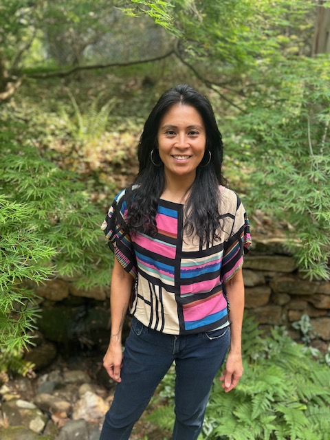 Woman standing outdoors among green trees and plants, smiling, wearing a colorful striped top and dark pants.