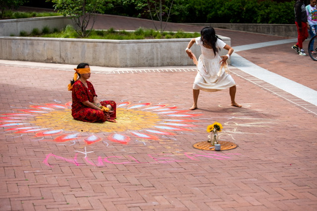 Child sitting on a colorful rangoli design on the ground, with a woman dancing nearby, in an outdoor area.