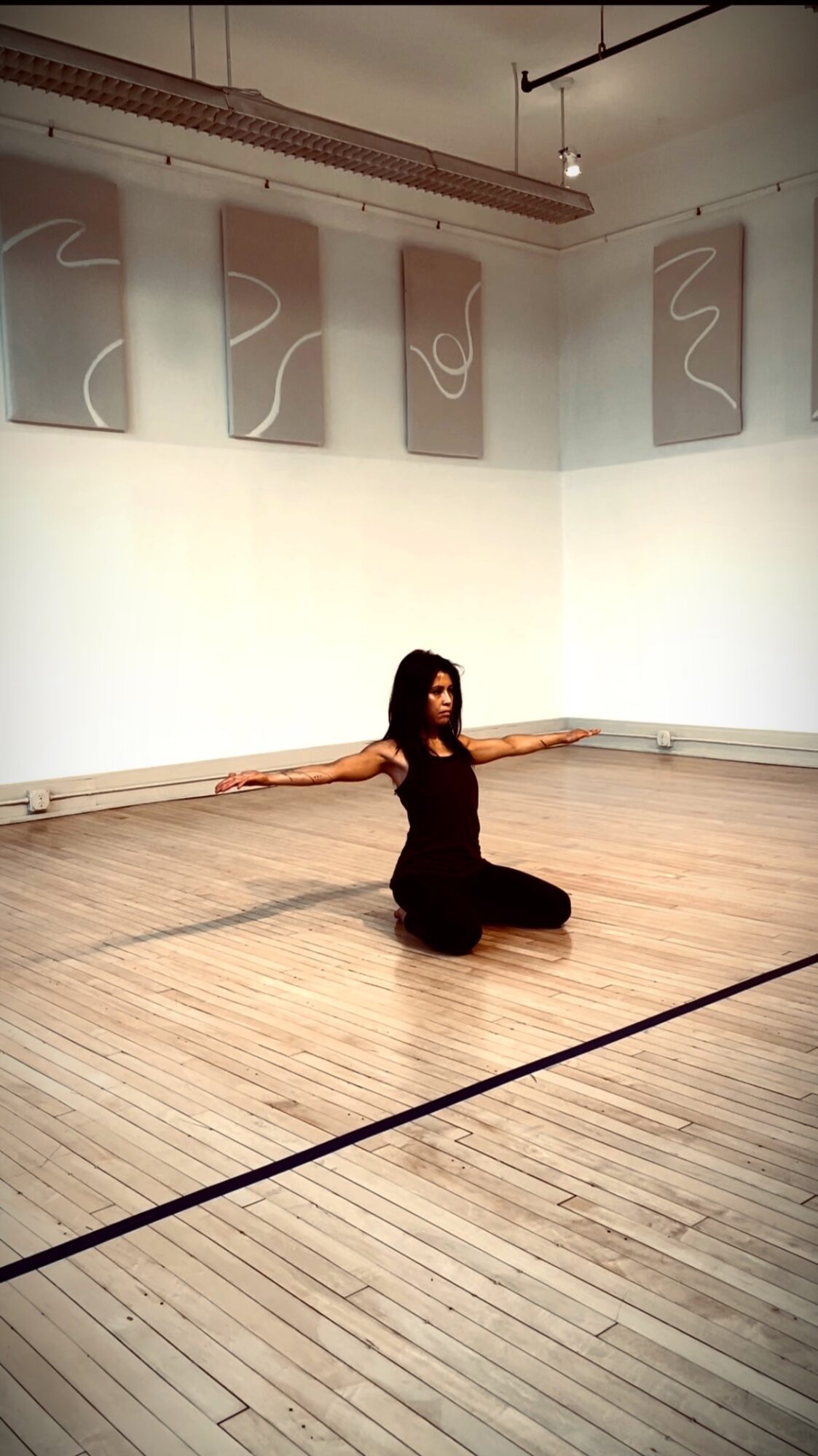 Person sitting on the floor with arms extended, in a room with abstract artwork on the walls and wooden flooring.
