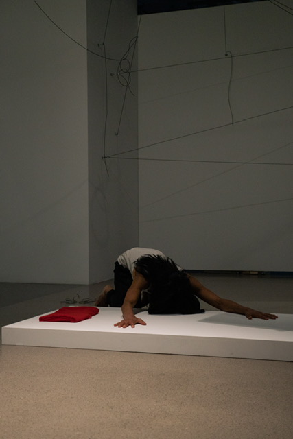 Person lying on a white platform with arms and legs extended, dark hair, white top, red object nearby, plain background.