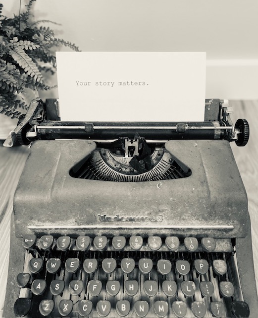 Typewriter with a card reading 'Your story matters.' on it, placed on a surface near a plant.