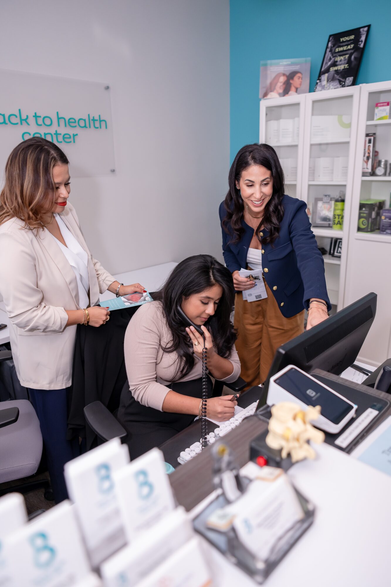 Three women in a health center, one sitting at a desk and two standing, smiling and talking. Medical supplies on desk.